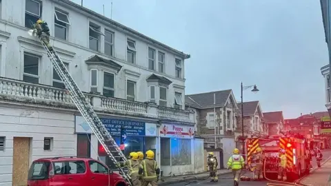 Island Echo Firefighters and fire engine in road below a large white building with a ladder against a third floor window and one firefighter looking in