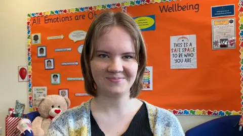 A young girl with short blonde hair smiles at the camera. She is standing in front of a red school notice board displaying information about emotions.