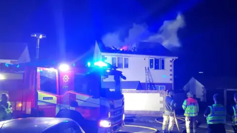 Flames and smoke rise from a collapsed roof on a house. It is night time and the scene is illuminated by the flashing blue lights of a fire engine parked in front of the two storey property.