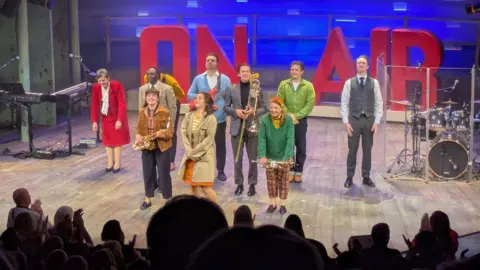 Lydia Geatrix At the end of a performance, nine actors prepare to bow. All are dressed in clothes from the 1960s and the words "on air" are in big red letters in the background