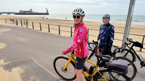 Lin Osborn saddling her bike and smiling at the camera. She is wearing a grey helmet, brown sunglasses, black cycling shorts and a red flowery cycling jacket. Behind her friend and a male cyclist wearing a black cycling shorts and blue cycling jacket. Poole Quay is in the background.