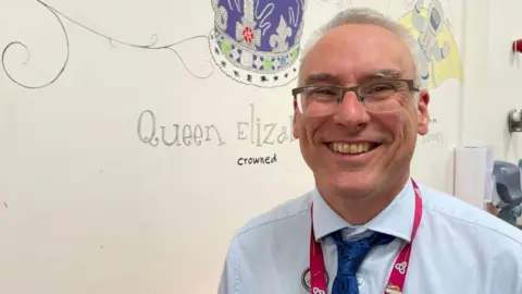 A man wearing glasses smiles in front of a library wall display which features the Crown of Queen Elizabeth II.