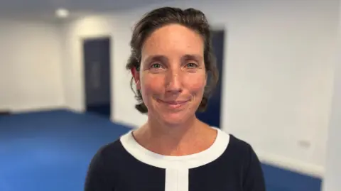 Deputy Lindsay de Sausmarez - A woman with medium length brown hair looking towards the camera. She's wearing a black blouse with a white collar and strip in the middle. 