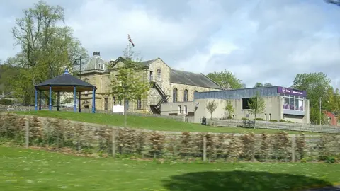 Stanley Howe A view across grass to the impressive Jedburgh Town Hall and a more modern addition of a low block that was once a tourist information centre. There is a shelter of some kind in front of them.