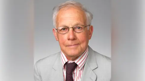 A portrait of a man with short white hair, glasses, a dark red spotted tie, a red and white shirt and a grey jacket.