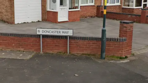 A picture of a street sign in a residential area that reads "Doncaster Way". The sign is in white and in front of a low red brick wall. There is a black lamp-post to the right of the sign.