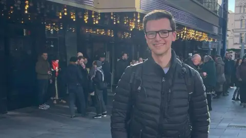 Dan Heard Dan, who has short dark hair and glasses and is wearing a black puffy jacket and black backpack. He is smiling and looking at the camera and behind him is the Odeon cinema in Leicester Square, with twinkling lights and crowds of people outside.