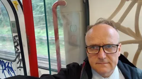 A man in glasses taking a selfie on the train with some unintelligible graffiti on the walls behind him