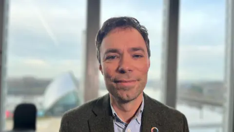 Rob Holland, a brown-haired man with a light stubble, smiles in a room with a window overlooking the Science Centre in Glasgow. It is a fair day behind him. He wears a brown tweed jacket and a light blue shirt.