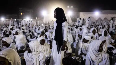 John Wessels/AFP Afbeelding van een jonge vrouw die voor een helder wit licht staat en een halo-achtig effect creëert, omringd door andere zittende vrouwen gekleed in geheel witte jurken in Dakar, Senegal - zondag 15 september 2024.