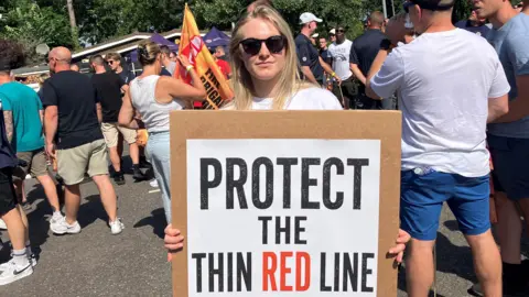 A woman with blonde hair holds a placard reading 'Protect the Thin Red Line'