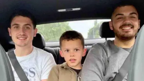Aime Hewitt Three brothers sit in the backseat of a car. All are smiling.