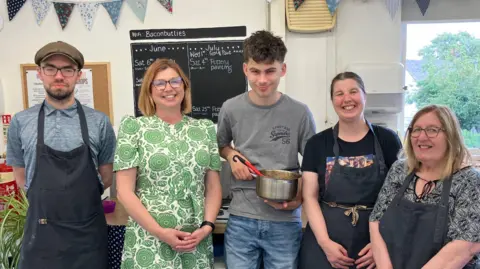 Five people stand in front of a blackboard. Jenny Lane who has re-started Workbridge wears a green and white patterned dress. A young man next to her holds a saucepan containing spagetti bolognaise.
