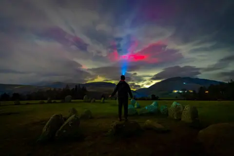 BBC Weather Watchers / Jonny Gios A person stands on one of the stones in the stone circle with his arms slightly outstretched. He is looking up at the night sky which has hues of pink and green behind the clouds.