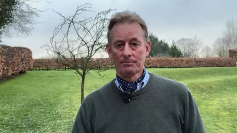 A man with light brown hair, a green jumper and a navy cravat is in front of a grassy lawn. Behind him is a browning hedgerow and a naked small tree.