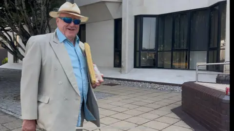 BBC Gwynoro Jones, pictured outside Swansea Crown Court. He wears a hat, sunglasses, a beige suit and a blue, open-collar shirt