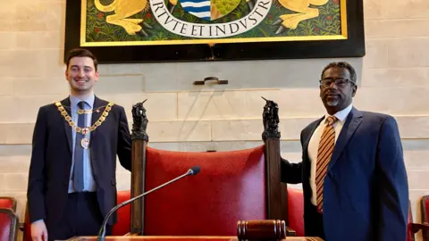 Henry Michallat, smiling, with his gold mayoral chains worn over his suit. He is standing to one side of a large red leather ceremonial chair in a council chamber, with Mohamud standing on the other side, also in a suit.