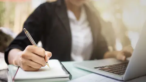 A female student takes notes in a pad as she types into a laptop computer.