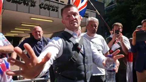 Tommy Robinson is wearing a light blue shirt and a checked blue waistcoat with a messenger bag over his shoulder. He is speaking with his hands open and outwards while holding a water bottle. A crowd of men surround him outside court.