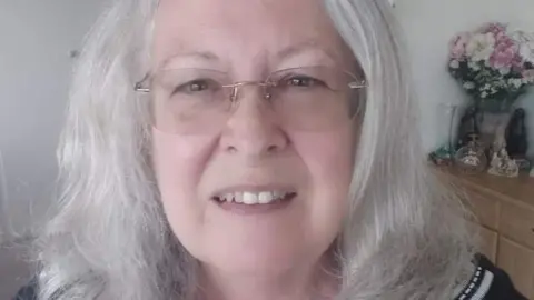 Pat Pollington Pat Pollington in glasses with a vase of flowers on a chest of drawers behind her.