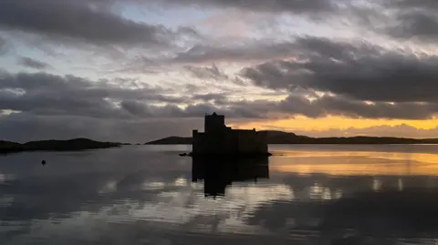 Maria Howat A dark castle completely in shadows at the centre of the image. The water below is calm and reflects the sky, which is grey and streaked with dark cloud. An orange glow from the setting sun is on the left side of the image.