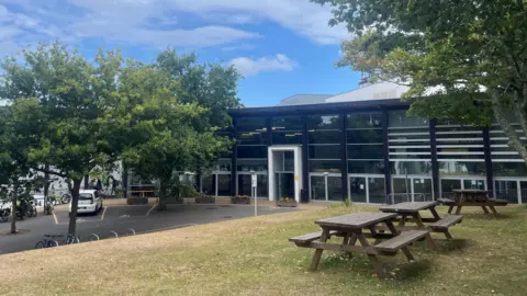 A black building, walled with dark glass. In the foreground is grass with 3 picnic tables on it. There are trees at either end of the picture.