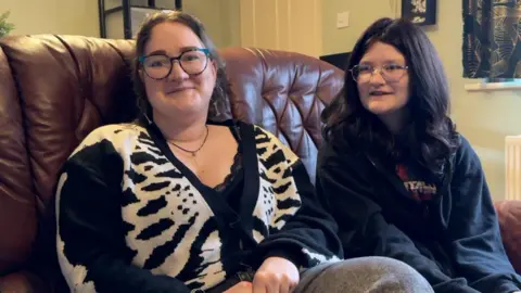 Coral Perkins-Bell and Alexa. Coral wears a black and white patterned cardigan and a black lace top. She has curly brown hair and glasses. Alexa has black hair with silver rimmed glasses and wears a black hoodie.