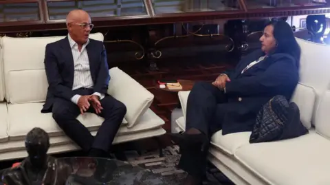 Jorge Rodríguez, wearing a dark suit and a white shirt and glasses, looks at Dinorah Figuera, who is wearing a navy suit, as they sit on white couches. 