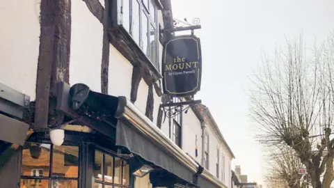 A white building with a thatched roof and many windows. A large black sign with the words The Mount can be seen hanging off the side of the building.