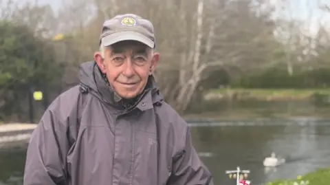 Head and shoulders of man in a grey hat and jacket stood looking at the camera with a canal in the background