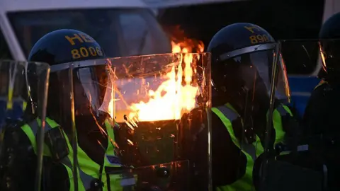 Getty Images A mid shot of police in protective riot uniform, including helmets and shields. There is a flaming bin in the background.