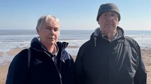 Kevin Shoesmith/BBC Gill Simpson dan suaminya Dennis, keduanya mengenakan jaket gelap, berdiri dengan punggung ke laut di Skegness Beach. Dennis mengenakan topi dan kacamata wol.