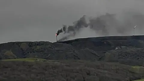 A slightly blurry photo of the wind turbine on fire in the distance on top of a grassy hill. Flames can be seen on the top of the white structure with black smoke coming off it. There is another wind turbine to the right which is not on fire.