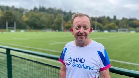 A man in an MND Scotland T Shirt stands smiling in front of a playing field in the Borders