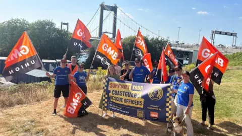 A number of people wearing blue t-shirts standing together holding range 'GMB Union' flags while smiling at the camera. 