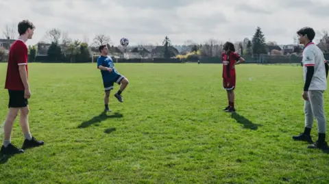 Bedfordshire Police Four boys standing on a football pitch in sports gear, kicking a football around together. There are houses and trees in the background.