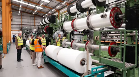 HFL/SCM Huge reels of yarn are seen stacked on spools to the right while on the left delegates examine them. 