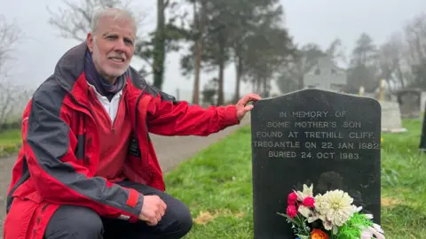 Simon Owens is wearing a red and black waterproof jacket and is looking at the camera, he is crouched beside a black gravestone with trees, grass and fog behind. The gravestone has yellow and pink flowers in front of it and reads: "In memory of Some Mother's Son, found at Trethill Cliff, Tregantle on 22 Jan 1982. Buried 24 October 1983."