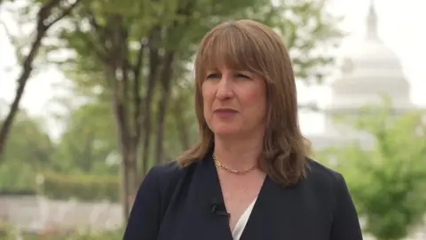 Head and shoulders of Rachel Reeves in dark suit with serious expression, and Capitol Building in the background out of focus.