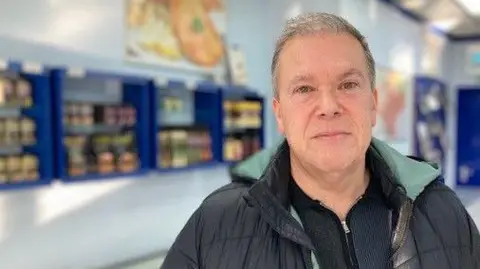 Michael has receding grey hair and blue eyes. He is wearing a black jacket over a black knitted top. He is standing in a shop. The background is out of focus, but shelves full of produce can be seen.