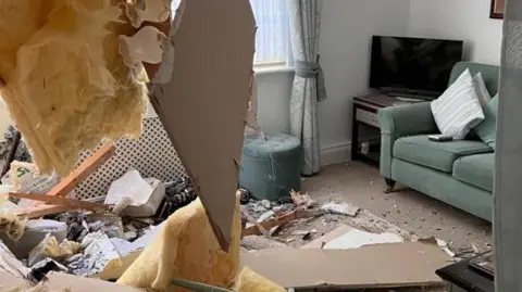 A normal living room with debris all over the carpet, insulation hangs down because the ceiling has fallen in 