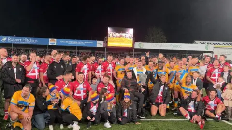 A mix of Rugby League players in yellow rugby shirts and Rugby Union players in red shirts on the pitch at Kingsholm Stadium. They're all stood together, celebrating as one of the rugby league players holds a silver trophy. Fans can be seen seated in the stands in the background as well as a big screen showing the match. It's night time and the sky is black. 