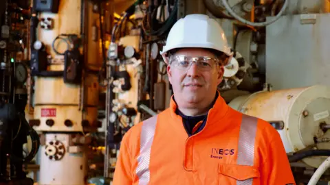 Ineos Energy Mads Gade, CEO of INEOS Energy, wears a bright orange, high-viz jacket, safety glasses and a white helmet. He stands in front of pipes and pumping equipment.