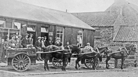 Beamish, The Living Museum of the North Historic black and white photograph, dating from the early 1900s, of three pony and carts. Two of the carts are behind the following pony, but the one of the left is visible and is labelled "pure ices". Behind is a group of about a dozen men wearing flat caps, and a long low building with a pitched roof, two doors and windows. The the right, can be seen the side of a larger stone building with a pitched tiled roof.