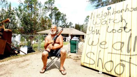 John Ferguson A man sits in shorts and flip flops, holding a gun. He is next to a sign which reads "neighbourhood watch you loot we all shoot".