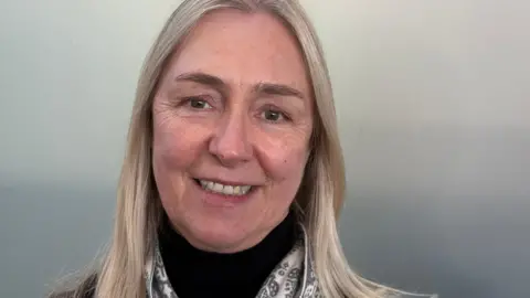 Amanda Kynaston Amanda Kynaston has shoulder length blonde hair and is wearing a black polo neck jumper with a light grey and cream scarf. She is smiling at the camera but the background is nondescript