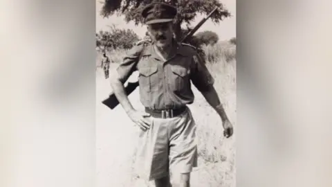 Family handout Jim Almonds, pictured in military issue shorts and short sleeves, is walking through grasslands. He has a rifle slung over his back.