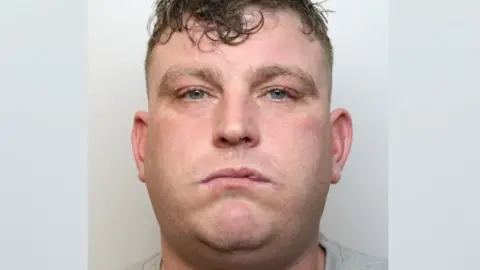 Wiltshire Police A mugshot of a man in a grey T-shirt looking seriously at the camera. He has brown hair, shaved at the sides and longer on top. It looks messy and wet. He has blue eyes and he looks tired. There is something on his lip, possibly a cold sore.