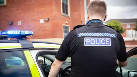 A police officer in a black top with the word police across his back is standing by the open door of a police car