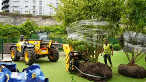 PA A yellow JCB vehicle deposits trees in the garden covered in cling film. A worker in a high vis stands between the plants. 